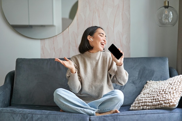 Portret van zorgeloos aziatisch meisje zit thuis, luistert naar muziek in koptelefoon en zingt op smartphone houdt mobiele telefoon vast als microfoon