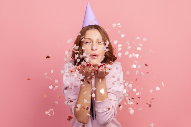 Portret van zorgeloos vrolijke tienermeisje in hoodie en partij kegel waait hartvormige confetti genieten van verjaardag of Valentijnsdag feestelijke stemming Indoor studio shot geïsoleerd op roze achtergrond