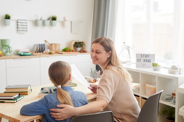 Portret van zorgzame lachende moeder meisje knuffelen zittend aan tafel en haar studie thuis helpen, kopie ruimte