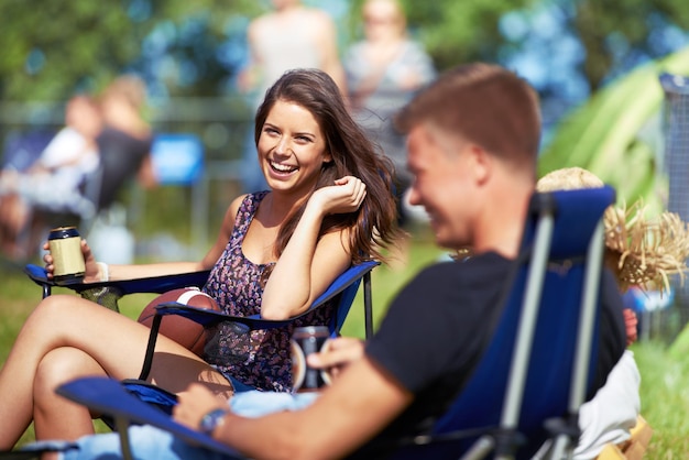 Portret vrouw die een biertje drinkt op een muziekfestival of camping en lacht met haar vriend vakantie gelukkige vrouwelijke persoon zittend op een kampeerstoel en een drankje drinkend terwijl ze met haar praat