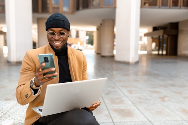 Portret zwarte zakenman in elegante stijlvolle kleding zittend op trappen in de stad en met behulp van computerlaptop. guy lacht tijdens het chatten met smartphone.