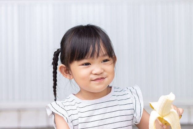 Portretbeeld van baby 3-4 jaar oud. gelukkig aziatisch kindmeisje geniet van het eten van een banaan met een zoete glimlach. voedsel en gezond kinderconcept.