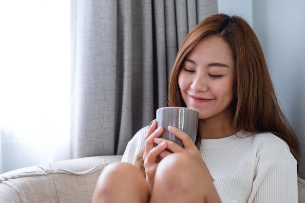 Portretbeeld van een mooie Aziatische vrouw die hete koffie drinkt en thuis op een bank ontspant