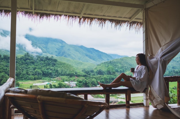 Portretbeeld van een mooie aziatische vrouw die hete koffie vasthoudt en drinkt op het balkon, kijkend naar bergen en groene natuur