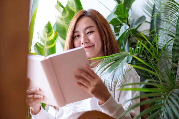 Foto portretbeeld van een mooie jonge aziatische vrouw die een boek leest op balkon met kamerplantentuin thuis