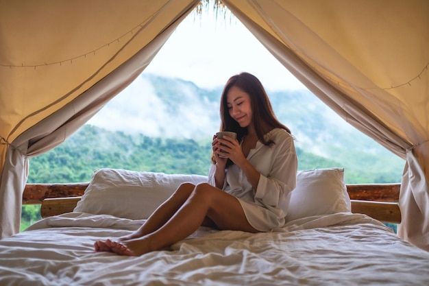 Portretbeeld van een mooie jonge Aziatische vrouw die koffie drinkt terwijl ze 's ochtends op een wit bed zit met een prachtig uitzicht op de natuur buiten de tent