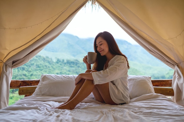 Portretbeeld van een mooie jonge Aziatische vrouw die koffie drinkt terwijl ze 's ochtends op een wit bed zit met een prachtig uitzicht op de natuur buiten de tent