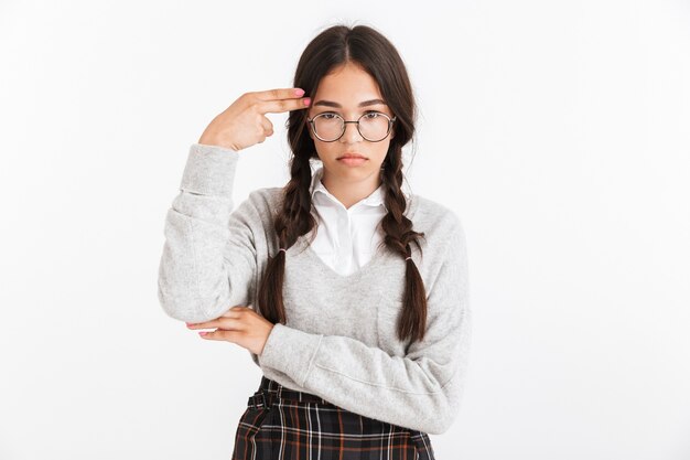 Portretclose-up van depressief tienermeisje die oogglazen en schooluniform dragen die het gebaar van het vingerkanon bij haar tempel tonen die over witte muur wordt geïsoleerd