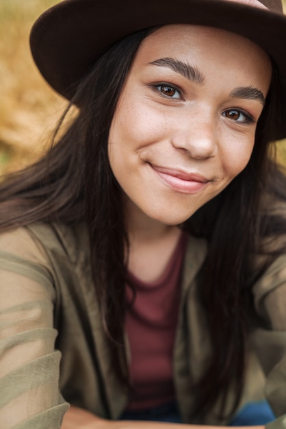 Portretclose-up van een schattige vrouw met lang donker haar met een hoed die naar de camera glimlacht terwijl ze selfie maakt