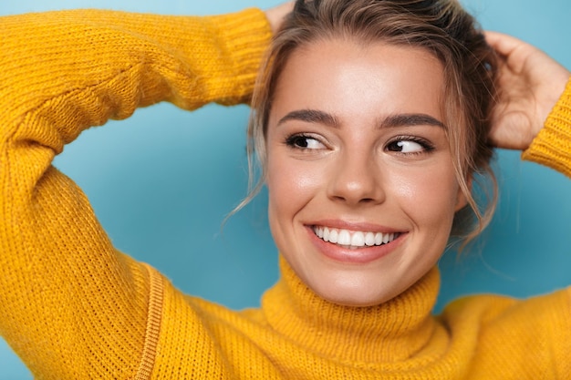 Portretclose-up van jonge tevreden vrouw in gele sweater die glimlacht en opzij kijkt geïsoleerd op blue