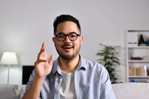 Portretfoto van een gelukkige, vriendelijke en zelfverzekerde Aziatische zakenman die thuis op een bank zit, naar de camera kijkt en praat en met de hand zwaait en hallo zegt tijdens een videogesprek