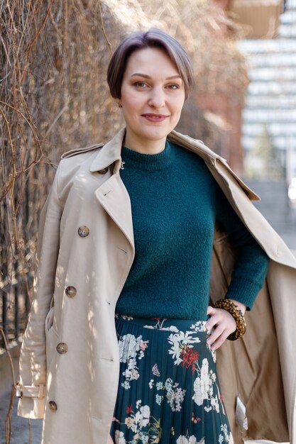 Portretfotografie van een aantrekkelijke, stijlvolle vrouw met een kortere weg, meisje loopt buiten in de stad. Stijlvol modern en vrouwelijk beeld, stijl. Meisje in een beige mantel of jas en een groene jurk