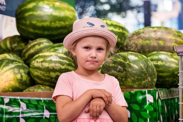 Portretmeisje dat in de fruitwinkel staat en diep nadenkt en naar de camera kijkt bij watermeloenenachtergronden