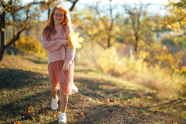Portretten van een charmant roodharig meisje met een schattig gezicht. Het stellen van het meisje in de herfstpark in een sweater en een koraalkleurige rok. In de handen van een meisje een geel blad