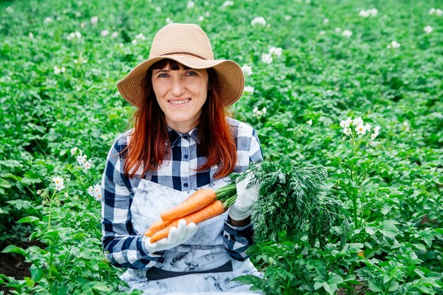 Portretvrouwenboer houdt een bos wortelen in een strohoed op de achtergrond van een moestuin