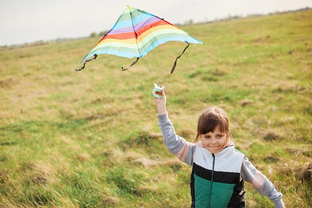 Portrsait of happy little girl flying a colorful kite running\
and jumping on a green meadow wearing coat spending day in active\
way having fun alone running happily
