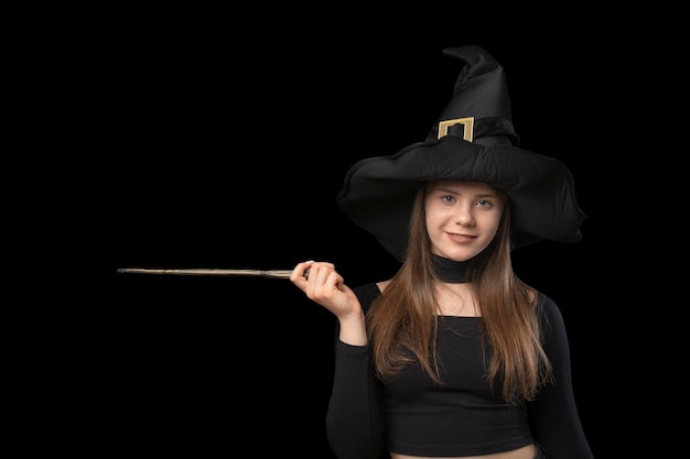 Porttrait of young witch in pointed hat Magic wand in hands Copy space for text Isolated on black background Halloween costume