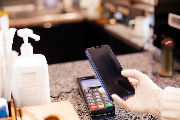 Pos-terminal bij café- of restaurantbalie Contactloze betaalmethode om sociale afstand te bewaren tijdens pandemie Close-up vrouw handen in beschermende handschoenen met bankkaart in de buurt van pos-terminal