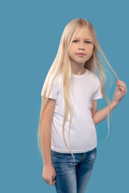 Poseren. Schattig kalm meisje in witte t-shirt en spijkerbroek die haar lange blonde haar aanraakt