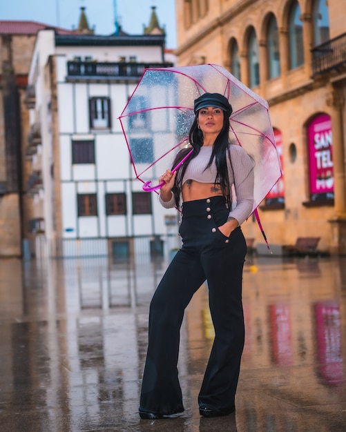 Poseren van een jonge brunette Latina met een leren hoed in de regen met een transparante paraplu in de stad