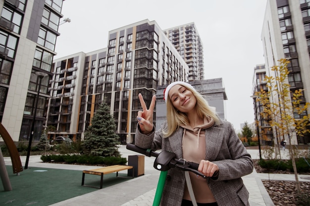 Poseren voor foto Jonge vrolijke blonde vrouw vredesteken tonen en camera kijken met een glimlach. Grijze moderne flatblokken op achtergrond. Ze rijdt op een elektrische scooter.