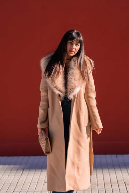 Posing of a brunette model in a winter jacket leaning against a wall with a bag. caucasian pretty a red background