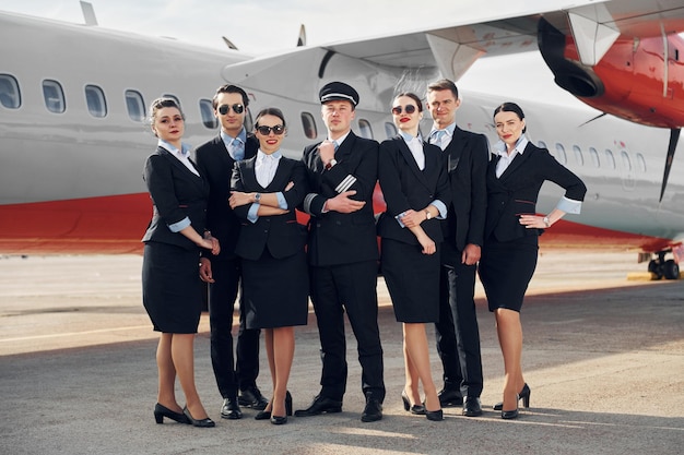 Foto in posa per una telecamera equipaggio di lavoratori aeroportuali e aerei in abiti formali in piedi insieme all'aperto