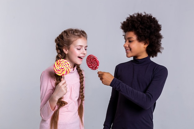 Posing for cameraman . Smiling dark-haired boy proposing his red candy to light-haired curious girl