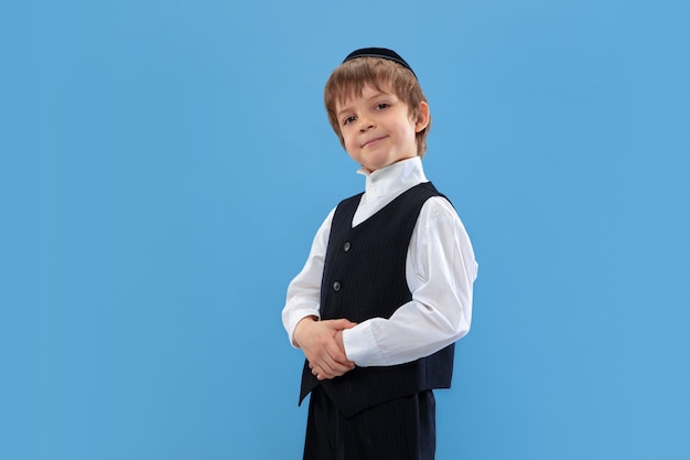 Posing confident, cute. Portrait of a young orthodox jewish boy isolated on blue wall. Purim, business, festival, holiday, celebration Pesach or Passover, judaism, religion concept.