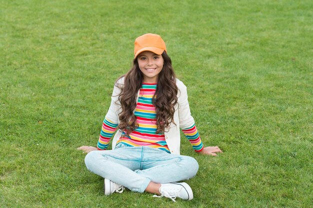 posing for photo. beautiful student teen girl in park. cute smiling confident schoolgirl relax on green grass. happy childhood. back to school. Small girl wear casual clothes. beauty and fashion.