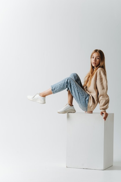 Posing for a photo teenage girl holding herself on a white cube. In a studio over white background.