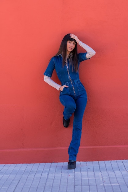 Posing of a smiling brunette girl leaning against a wall in a blue denim outfit. Pretty Caucasian posing on a red background