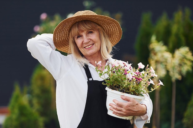 Posing with pot of flowers in hands Senior woman is in the garden at daytime Conception of plants and seasons