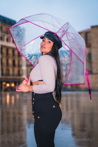 Posing of a young brunette Latina with a leather cap in the rain of the city with a transparent umbrella