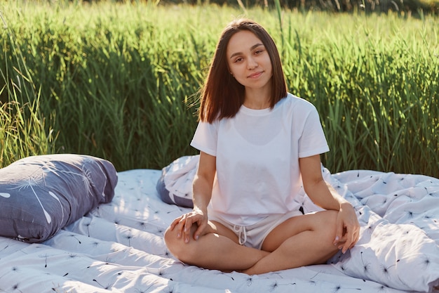 Positief aantrekkelijk meisje kleedt wit t-shirt en korte broek zittend met gekruiste benen op bed in groen veld in warme zonnige dag op zonsondergang