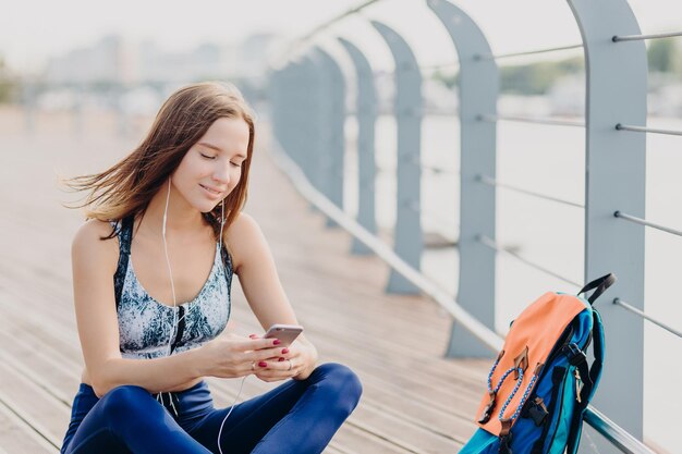 Positief fitnessmeisje in top en legging heeft een blije uitdrukking als een aangenaam sms-bericht dat van vriend is ontvangen, heeft donker haar rust buiten Mensen sporten moderne technologieën en levensstijl