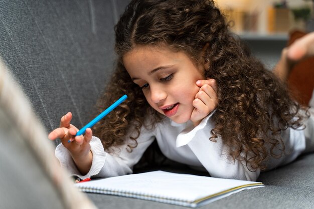 Positief geïnteresseerd krullend kind dat op de bank ligt en op haar notitieboekje schrijft terwijl ze huiswerk doet in de kinderkamer Stockfoto