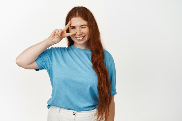 Positief gembermeisje met lang natuurlijk haar en sproeten witte glimlach die en vrede vsign tonen dichtbij gezichtsoog die zich in t-shirt tegen witte achtergrond bevinden