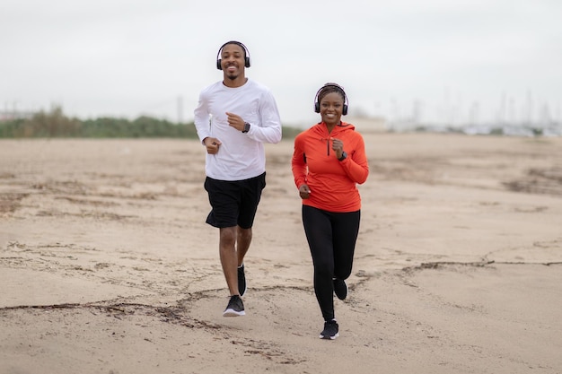 Positief jong afrikaans amerikaans stel in sportkleding en koptelefoons die samen op het strand rennen