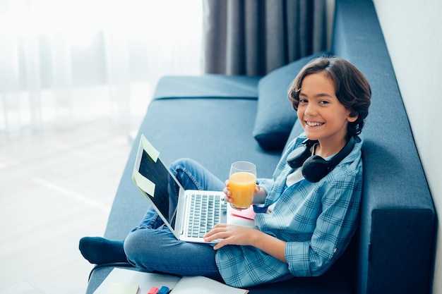 Positief kind dat thuis op een bank zit met een laptop op schoot en glimlacht terwijl hij een glas sinaasappelsap vasthoudt