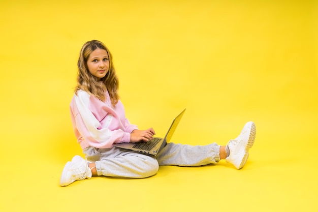 Positief kind zit met laptop, leest huiswerk en draagt een casual stijldoek geïsoleerd op een gele achtergrond