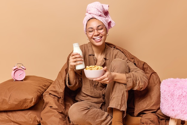 Positief lachende jonge vrouw draagt slaappak eet cornflakes met melk voor het ontbijt glimlacht en blijft gelukkig in bed in een goed humeur poseert tegen een beige achtergrond. Blij dat vrouwtje geniet van de ochtend