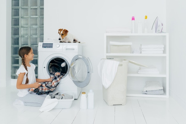 Positief meisje dat de wasmachine leegt, houdt een schoon geruit hemd vast en kijkt met een glimlach naar het favoriete huisdier dat helpt bij het doen van de was poses op een witte vloer met een bekken vol met schoonmaakmiddelen voor kleding