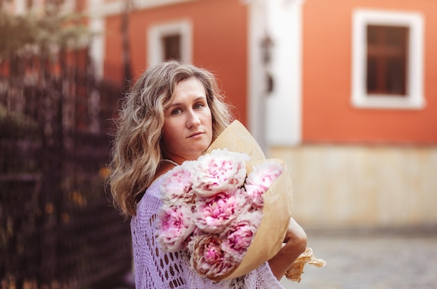 Positief meisje houdt bloemen in het centrum van de stad Close-up shot van geweldige blonde dame met natuurlijke make-up look gelukkig wandelen buiten houden roze boeket