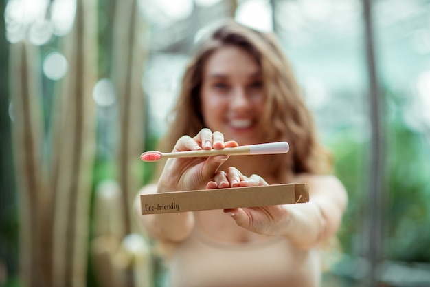 Positief meisje. Vrij jonge vrouw in beige kleren die een tandenborstel tonen en glimlachen