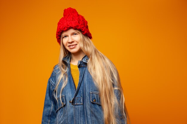 Positief millennial hipster vrouwelijk model in spijkerjasje en rode gebreide muts die naar de camera kijkt tegen een feloranje achtergrond