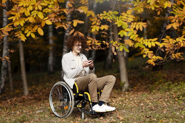 Positieve aantrekkelijke man met bril zit in een rolstoel en houdt een smartphone in zijn hand een gehandicapte jongen op straat onder een boom met gele bladeren