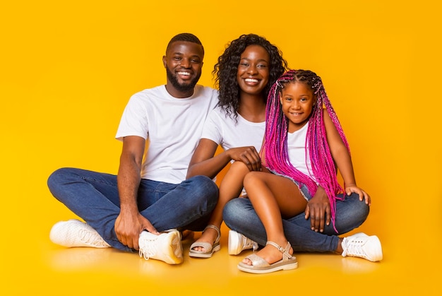 Positieve afro-amerikaanse familie van drie poseren voor de camera terwijl ze op de vloer zitten over een gele studioachtergrond, vrije ruimte