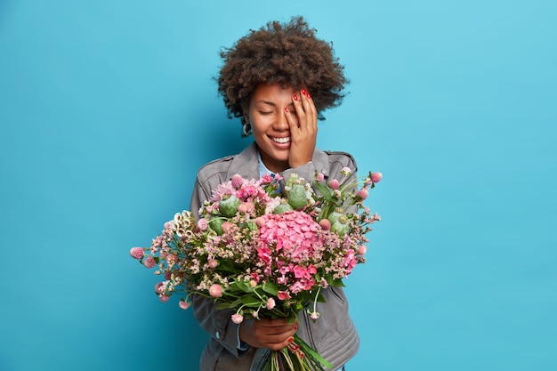 Positieve Afro-Amerikaanse vrouw houdt mooi boeket bloemen ontvangen op verjaardag covers gezicht met hand gekleed in grijze jas geïsoleerd over blauwe muur