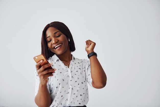 Positieve Afro-Amerikaanse vrouw in wit overhemd die binnen tegen een witte muur staat met telefoon in de hand